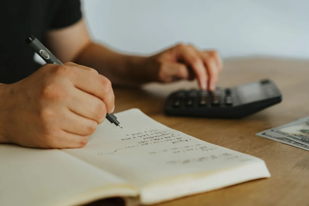 Hands writing calculations in a notebook beside a calculator