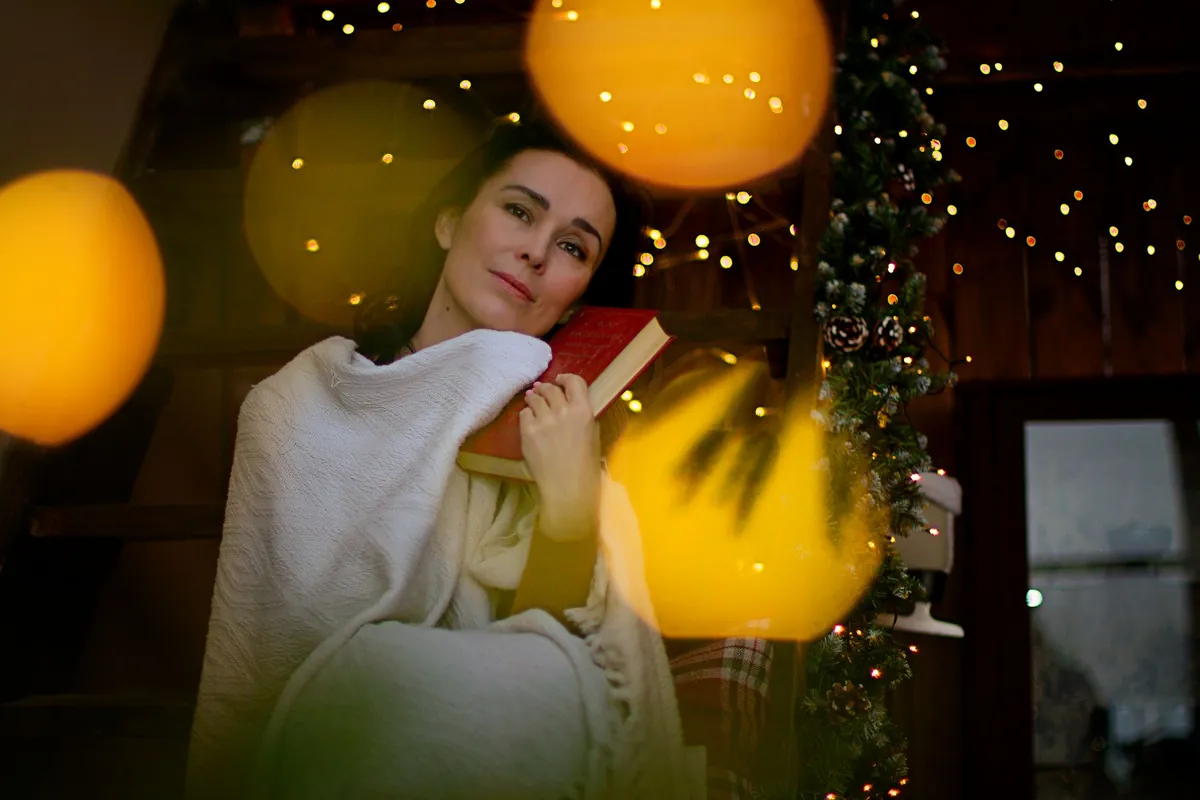 Woman wrapped in a blanket reading a book by holiday lights
